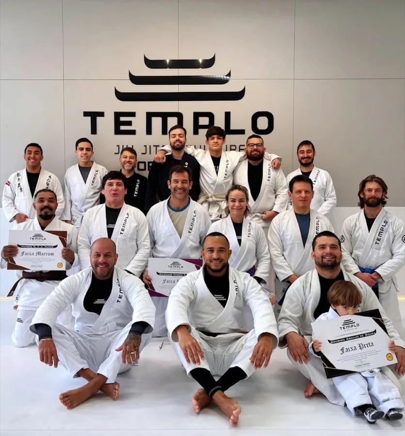 Turma da Templo Jiu Jitsu reunida no tatame sob o logo da academia, adultos e crianças de kimono, alguns segurando diplomas de graduação