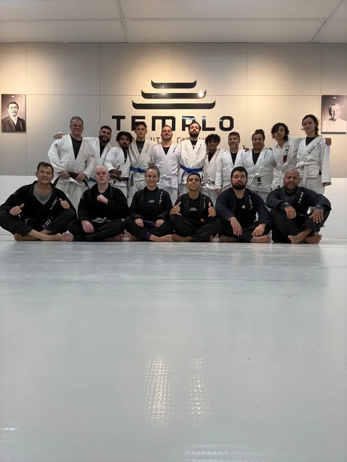 Turma adulta da Templo reunida no tatame em frente ao logo da academia, alunos de kimono branco e preto